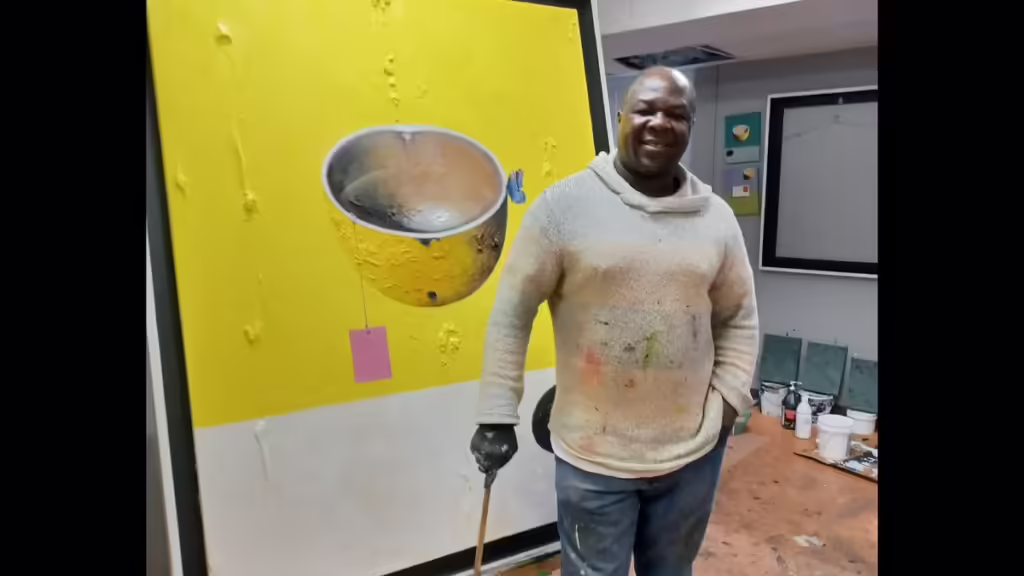 A person in a white hoodie that has a few paint stains on it stands smiling in front of a large yellow canvas that includes a painting of a silver bowl and butterfly.
