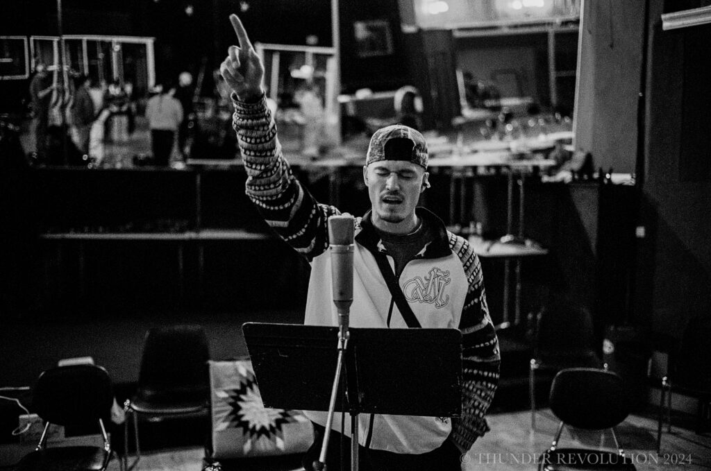 A person singing into a microphone, eyes closed. They have one of their arms raised and a finger pointing up.