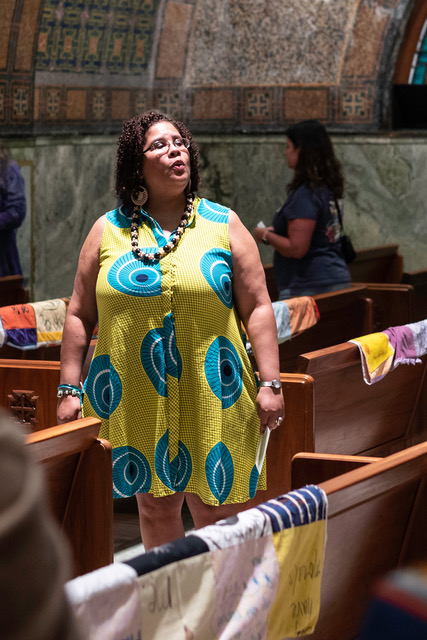 A person with medium dark skin tone wears a bright green dress and sings between pews indoors.