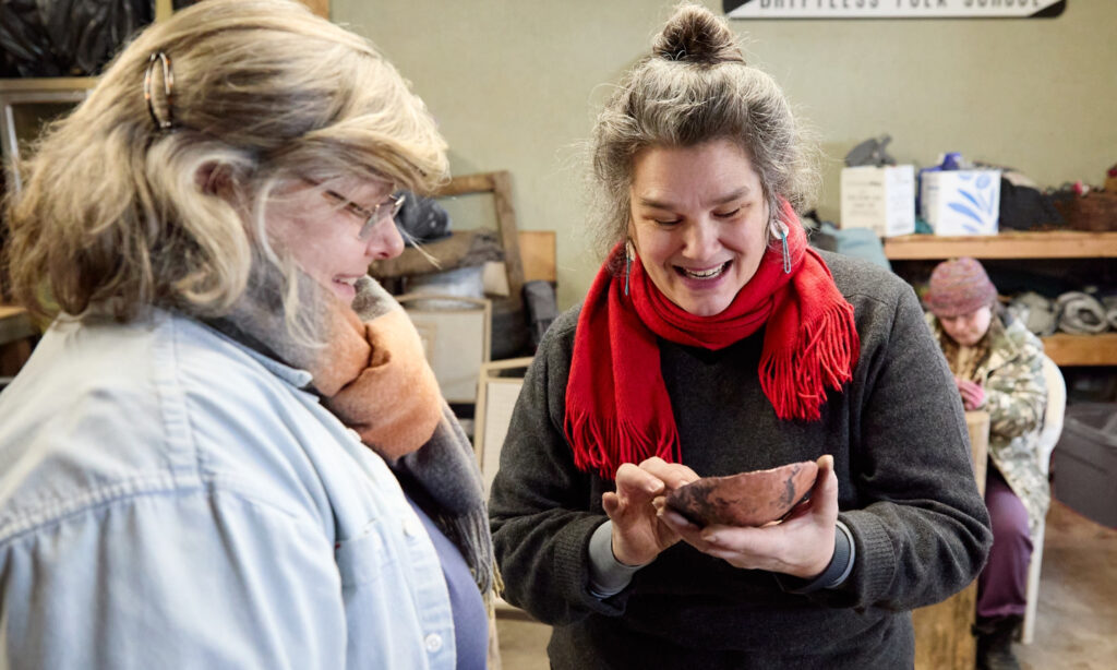 A person of medium-light skin tone with silver hair showing an adult student a copper bowl.