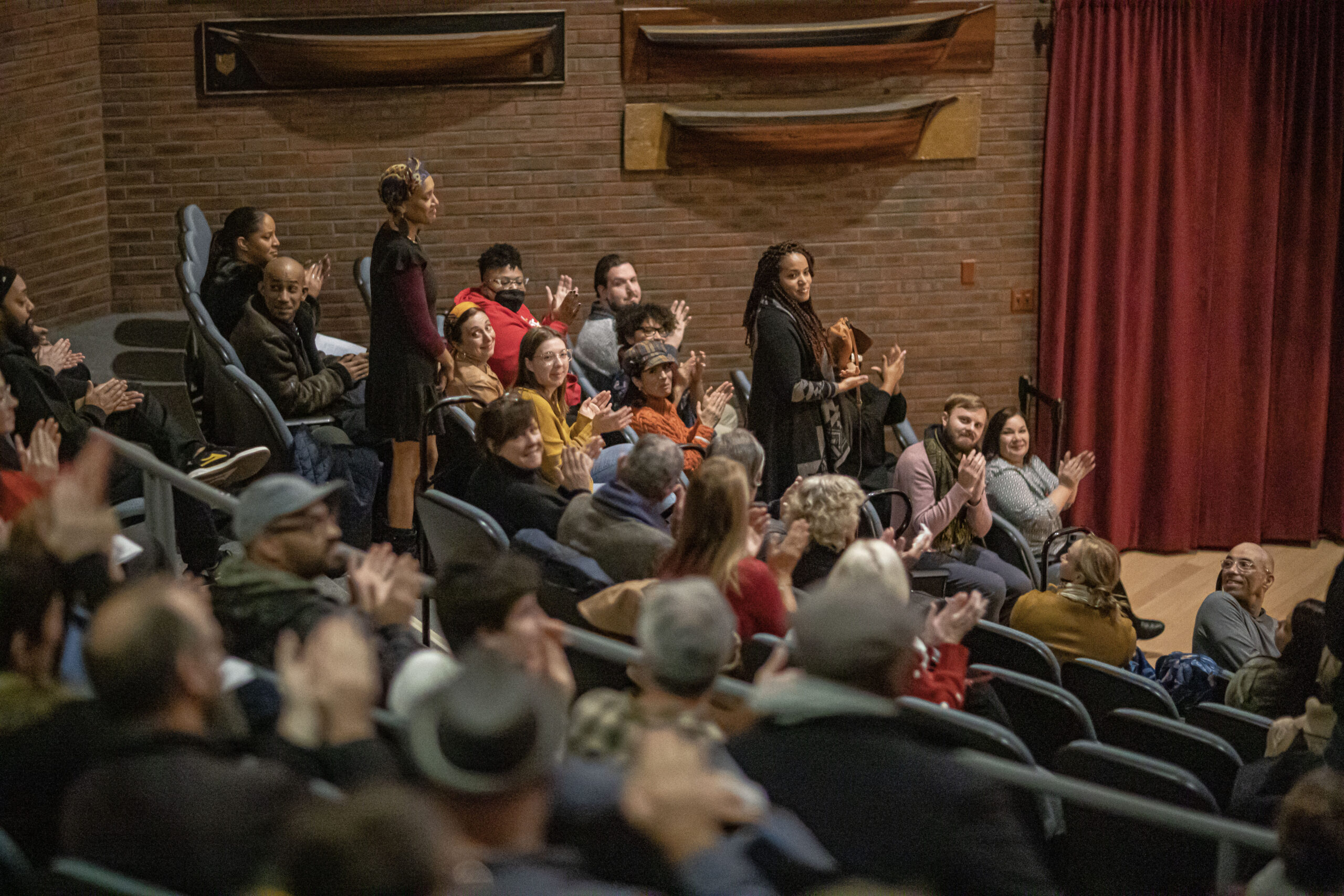 A crowd seated in a theater applaud as two people stand.