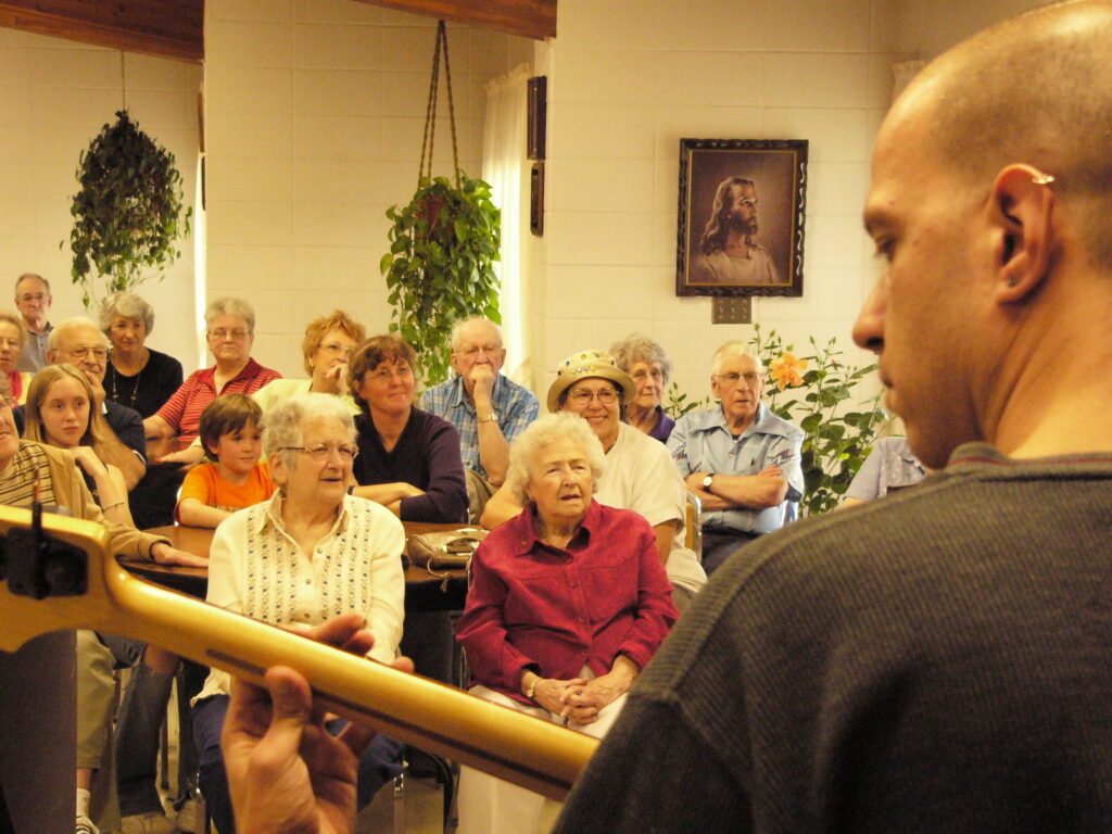 A musician plays bass guitar for an attentive audience of older adults seated in a cozy community room decorated with hanging plants and a portrait of Jesus.