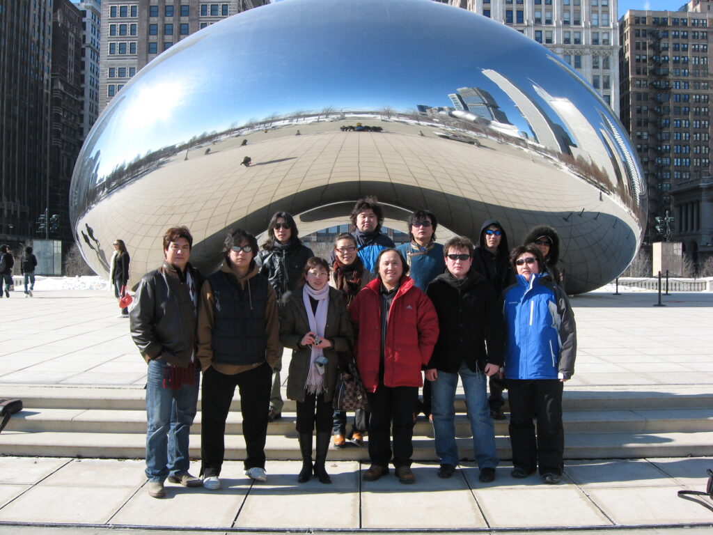 A group of performers in winter jackets pose in front of Chicago’s Cloud Gate sculpture, also known as “The Bean,” during a bright but cold day