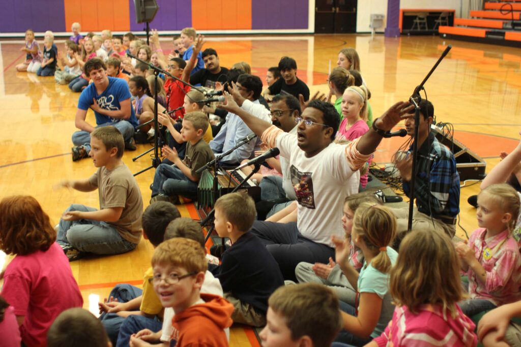 A group of musicians sits on the floor of a school gymnasium surrounded by children, enthusiastically clapping and participating in a musical workshop