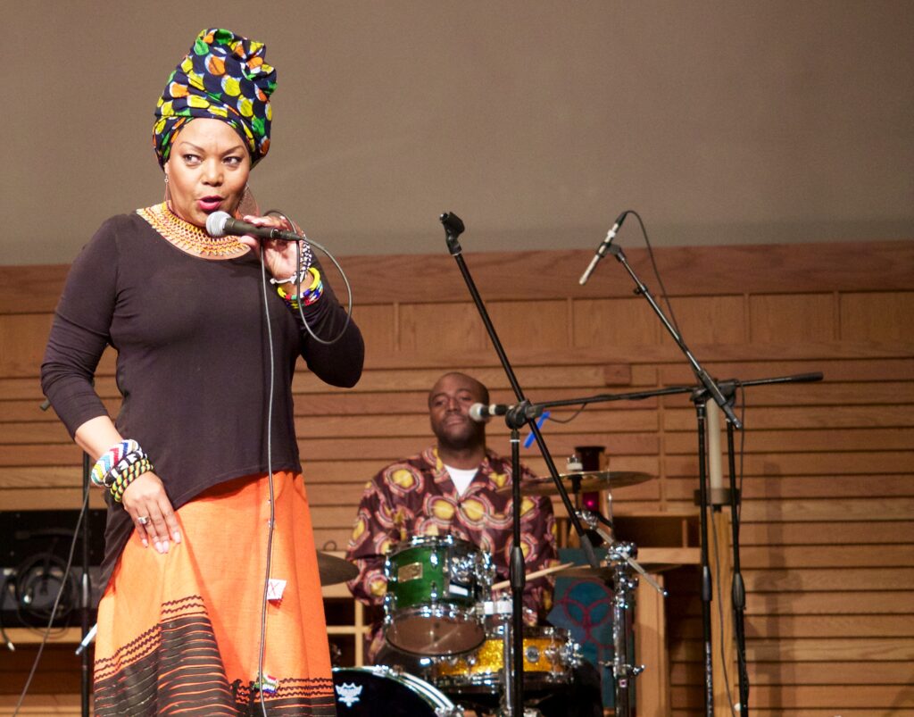 A vocalist in a vibrant headwrap and orange skirt sings into a microphone while a drummer plays behind her in a warmly lit performance space