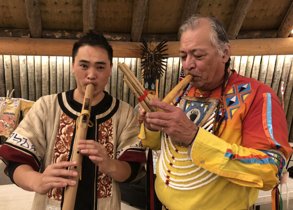 Two men wearing traditional clothing play wooden flutes together in a warmly lit room with wood-paneled walls and Native art in the background