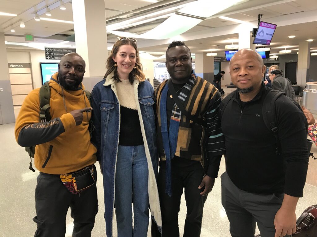 Four people pose together smiling at an airport. One man points playfully at the woman beside him, and everyone appears cheerful and relaxed.
