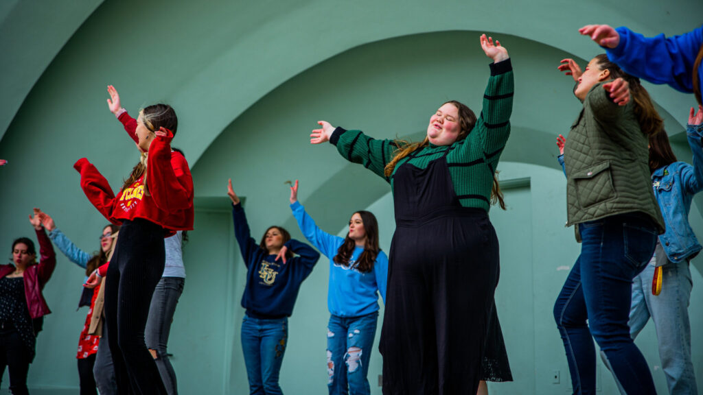 A group of teenagers dances with arms raised in front of a mint-green band shell, smiling and swaying in unison during a joyful outdoor performance.