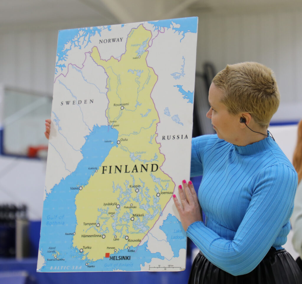 A performer in a bright blue shirt holds up a large map of Finland while speaking to a student audience in a gymnasium.