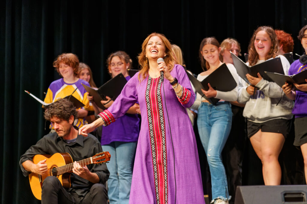 A vocalist in a flowing purple dress sings with joy while a guitarist plays seated beside her, accompanied by a student choir in the background.