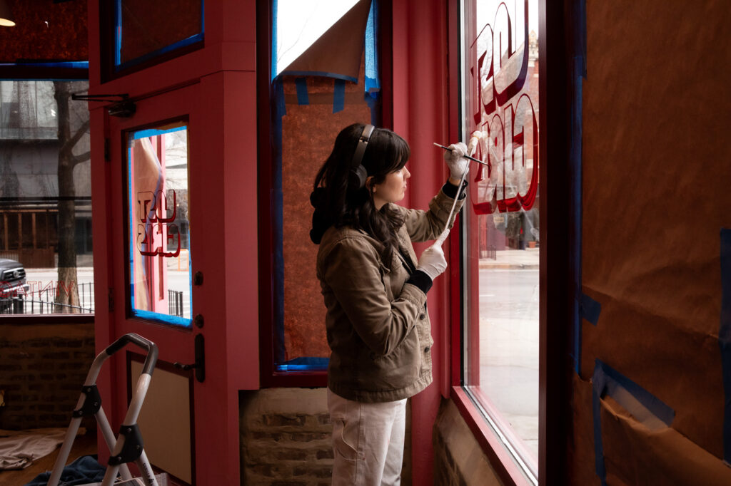 A person with light skin tone stands and paints a window indoors.