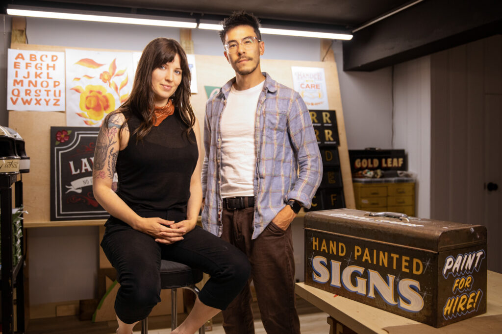 In an art studio under fluorescent lighting, a person with a light skin tone sits in a stool next to a standing person with medium-light skin tone.