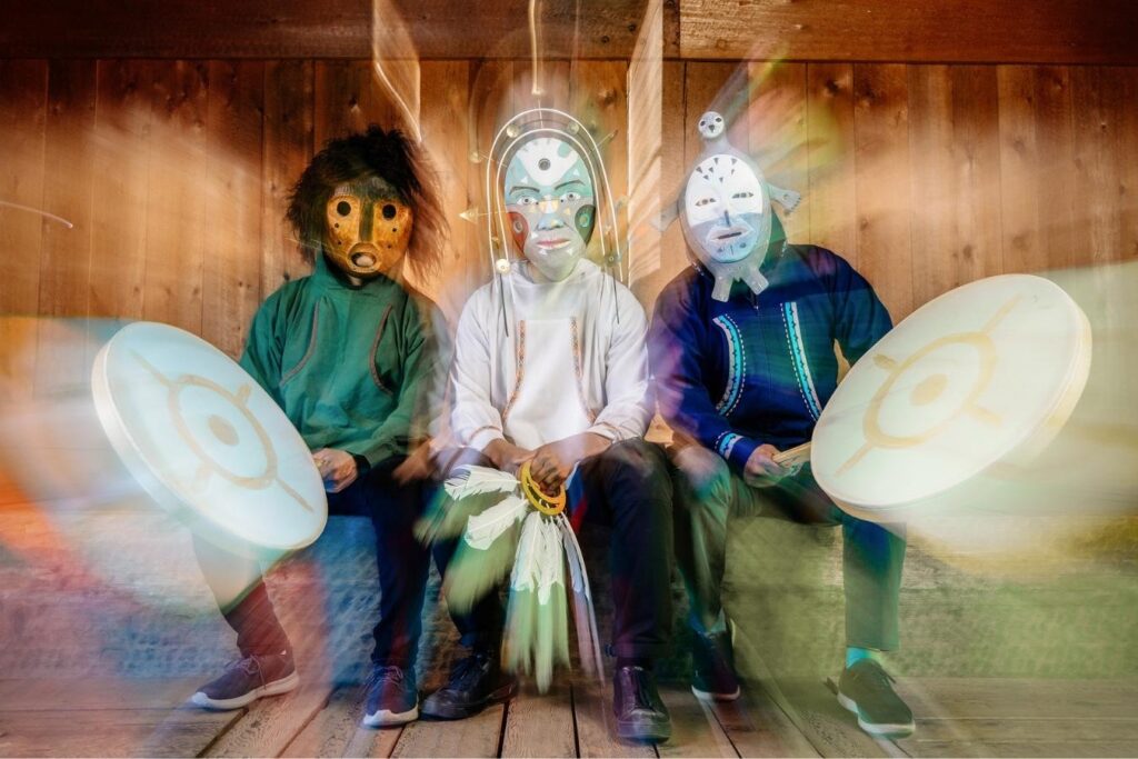 Yup’ik ensemble Pamyua sit together on a wooden bench, wearing traditional Yup’ik masks.