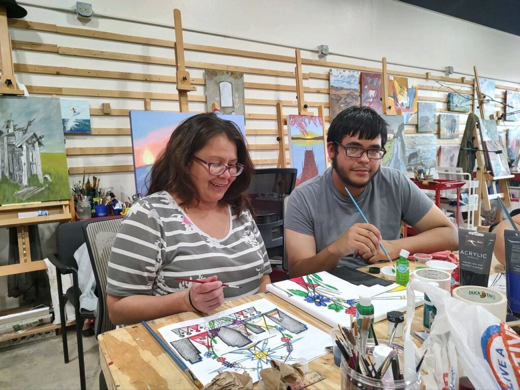 A woman and a man of medium skin tone paint canvases surrounded by art supplies