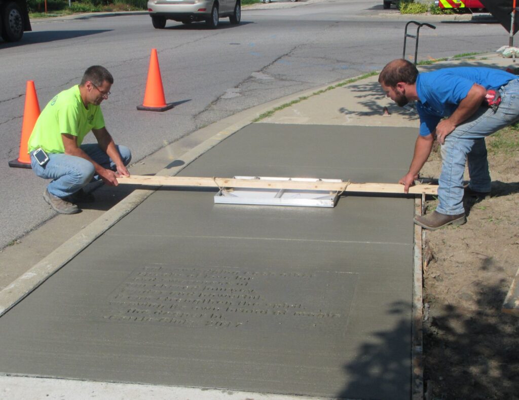 Two light-skinned people wearing construction gear place a piece of wood over a stamp on the sidewalk.