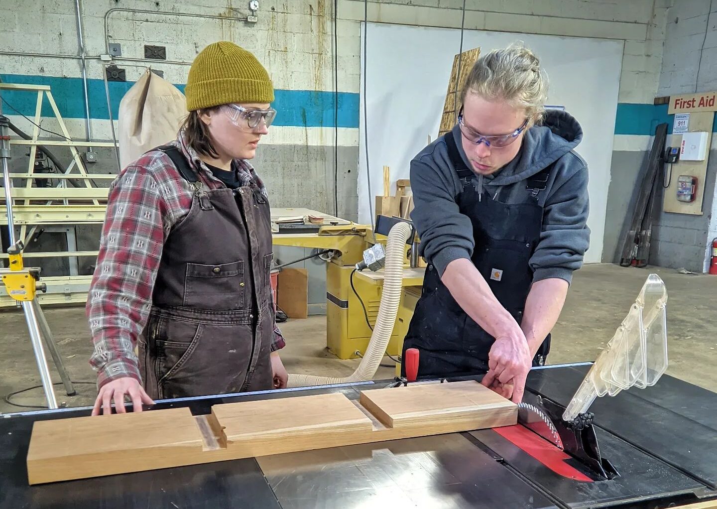 Two people wearing safety glasses and cutting a large piece of wood on a work table