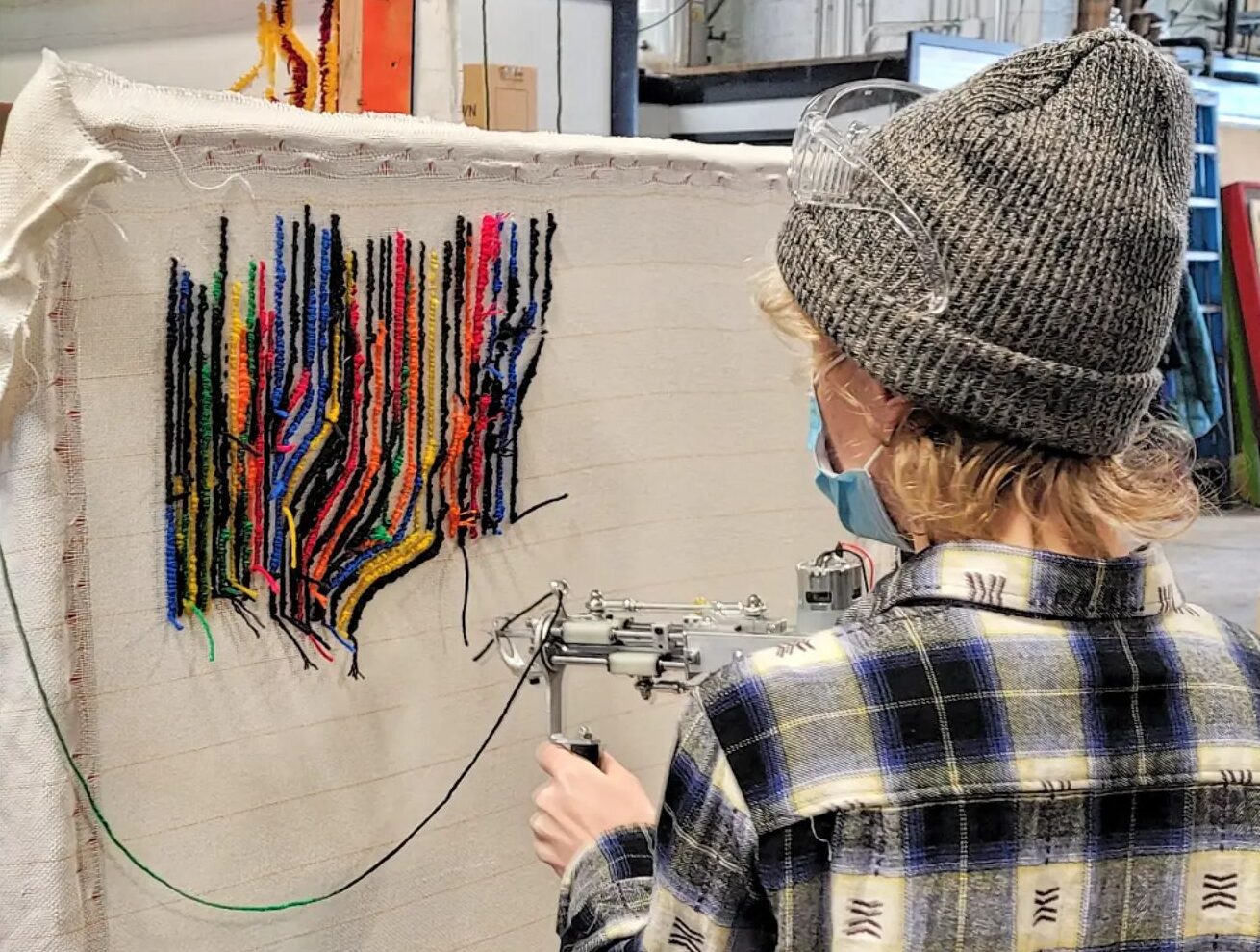 A person with blonde hair and light skin using a tufting gun and colorful material on a white stretched canvas.