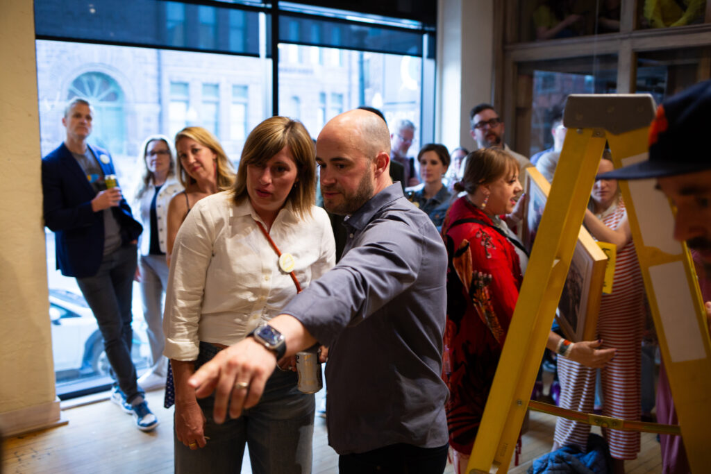 Two people standing next to each other. One of them is pointing to something. There are other people standing around and behind them.