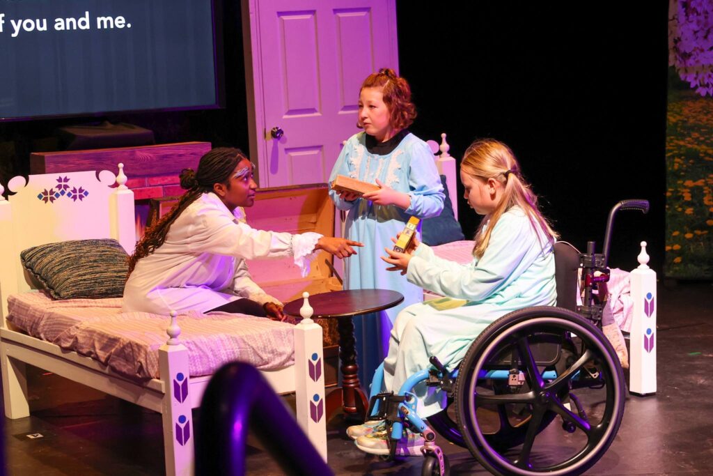 Three people act on stage near a bed prop.