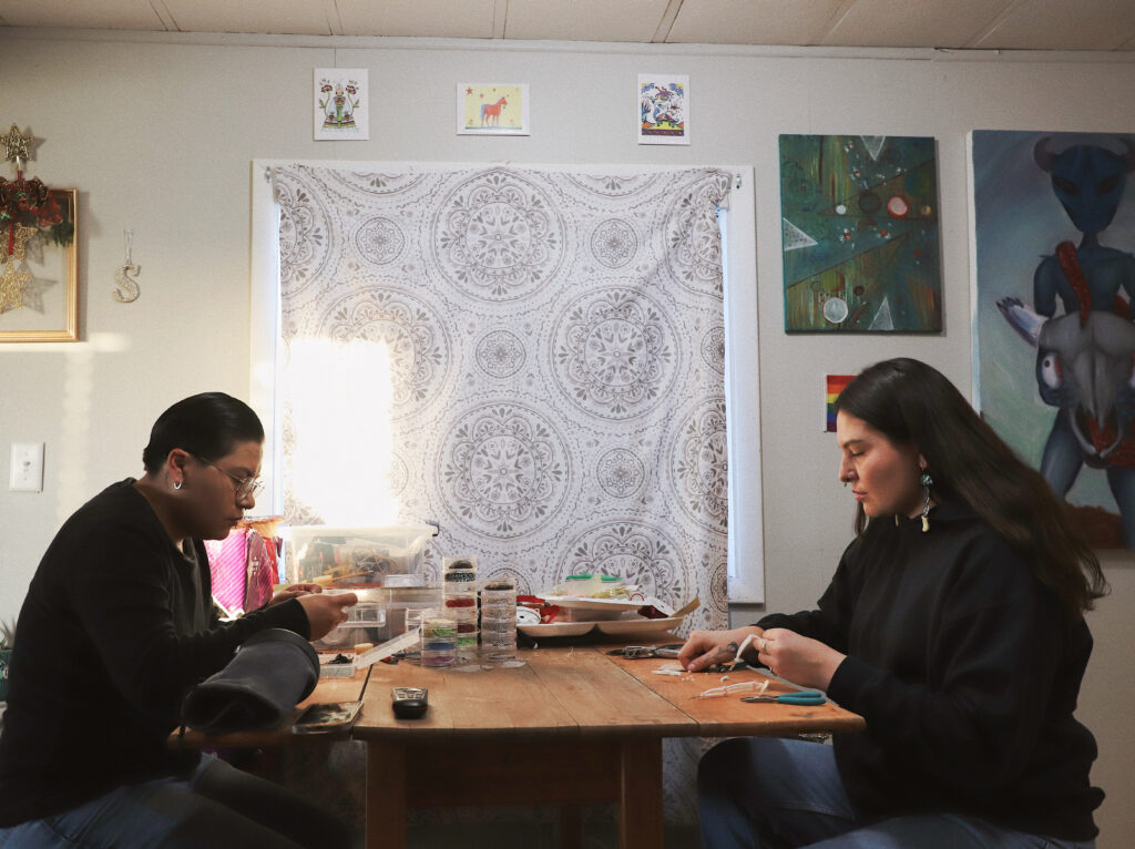 Two people of medium skin tone sit across from each other at a wooden table, heads down and working with their hands. The table has containers filled with materials and tools for beadwork and quillwork.