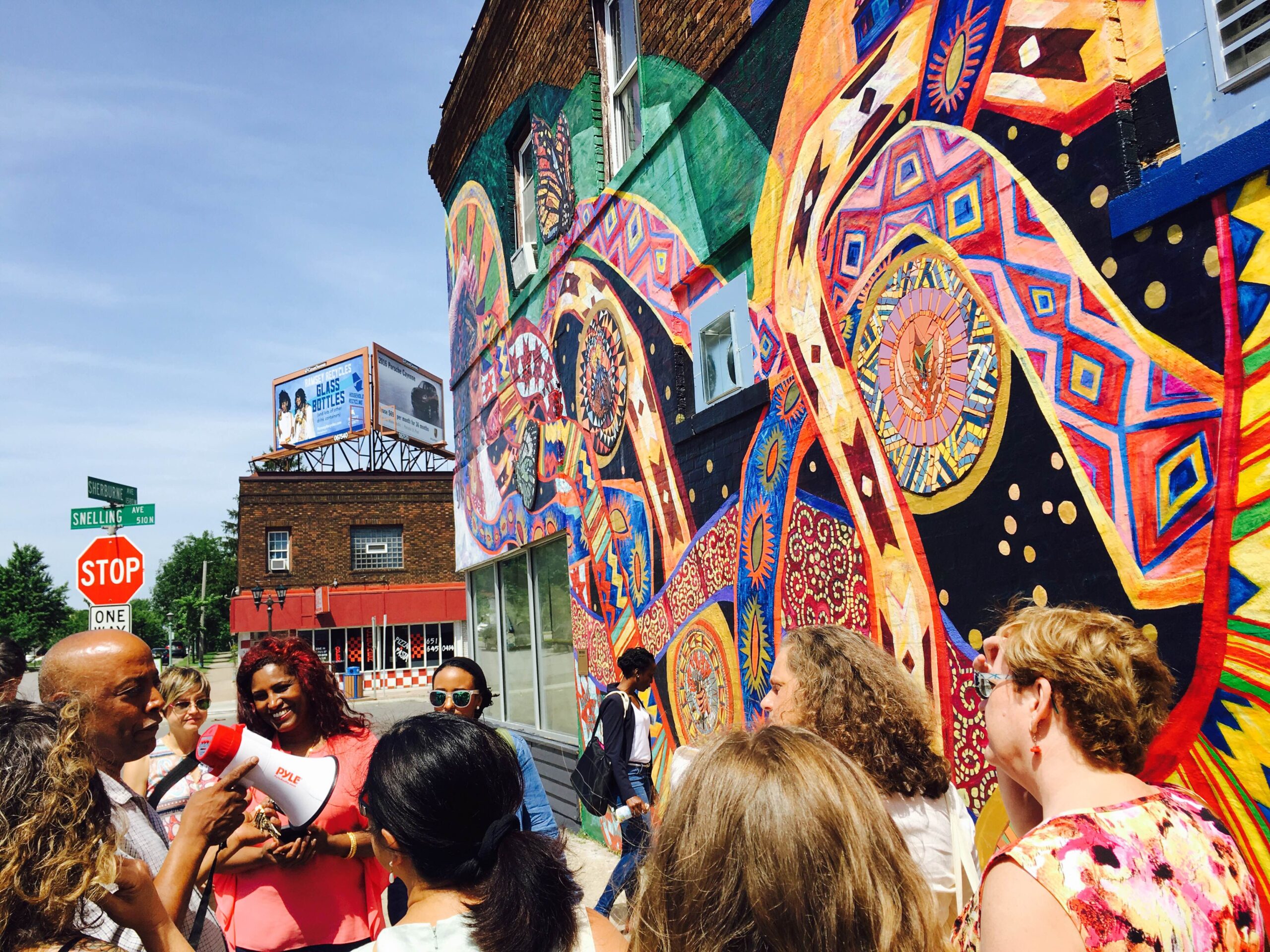 A person holding a megaphone speaks to a group of people standing outside a building with a colorful mural.