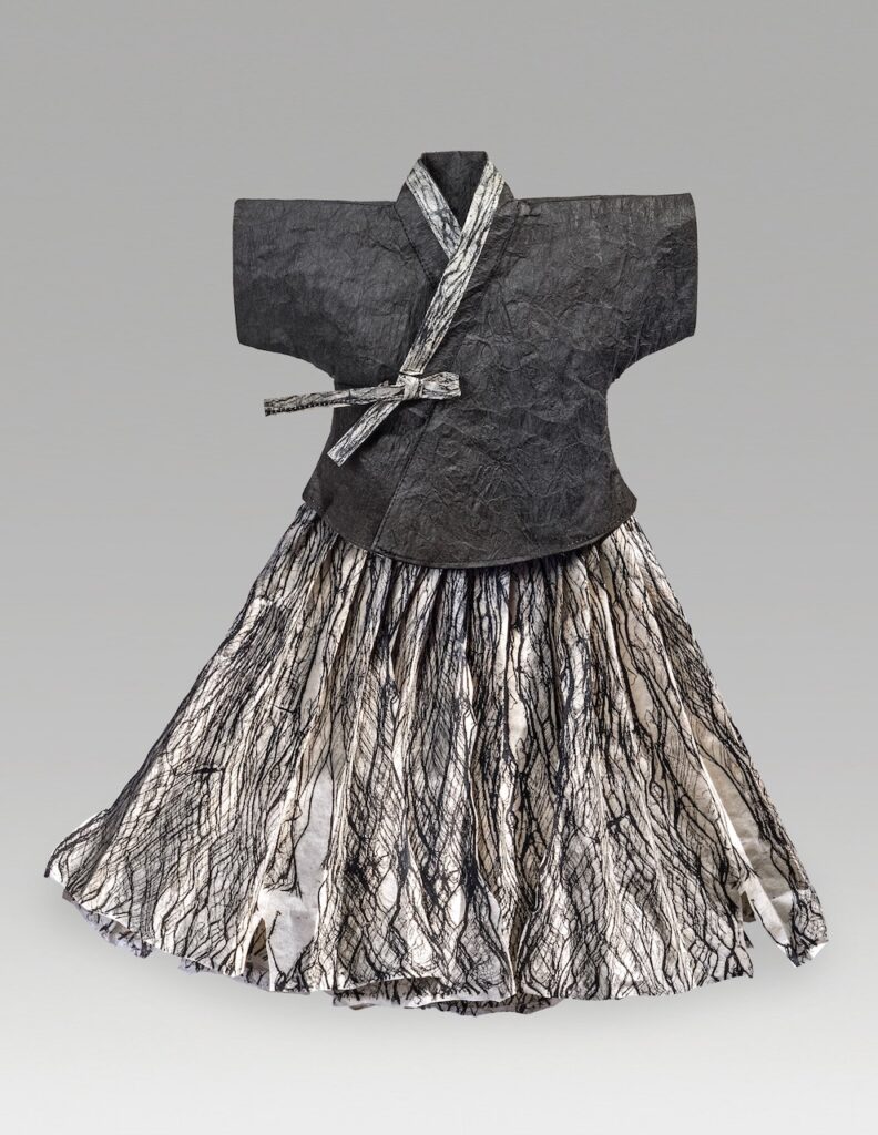 A dress made out of paper with a black short sleeved jacket and pleated skirt with black ink prints of laced bark on white paper.