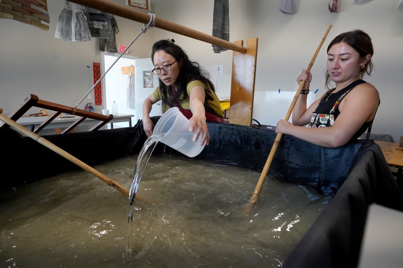 A person with long black hair pours gooey liquid into a large vat filled with fiber and water as a person with brown hair beside her agitates the vat with a long bamboo pole, inside of a papermaking studio.