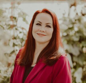 A smiling person of light skin tone with a reddish tint to their hair, wearing a magenta coat.