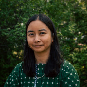 A woman with long, straight dark hair stands in front of a leafy green background. She is wearing a green checkered top with white embroidery, a silver necklace, and hoop earrings. She has a calm expression and a small nose stud.