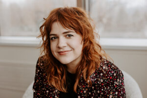 A smiling person of light skin tone with shoulder length wavy red hair, wearing a black blazer with multi-colored flowers