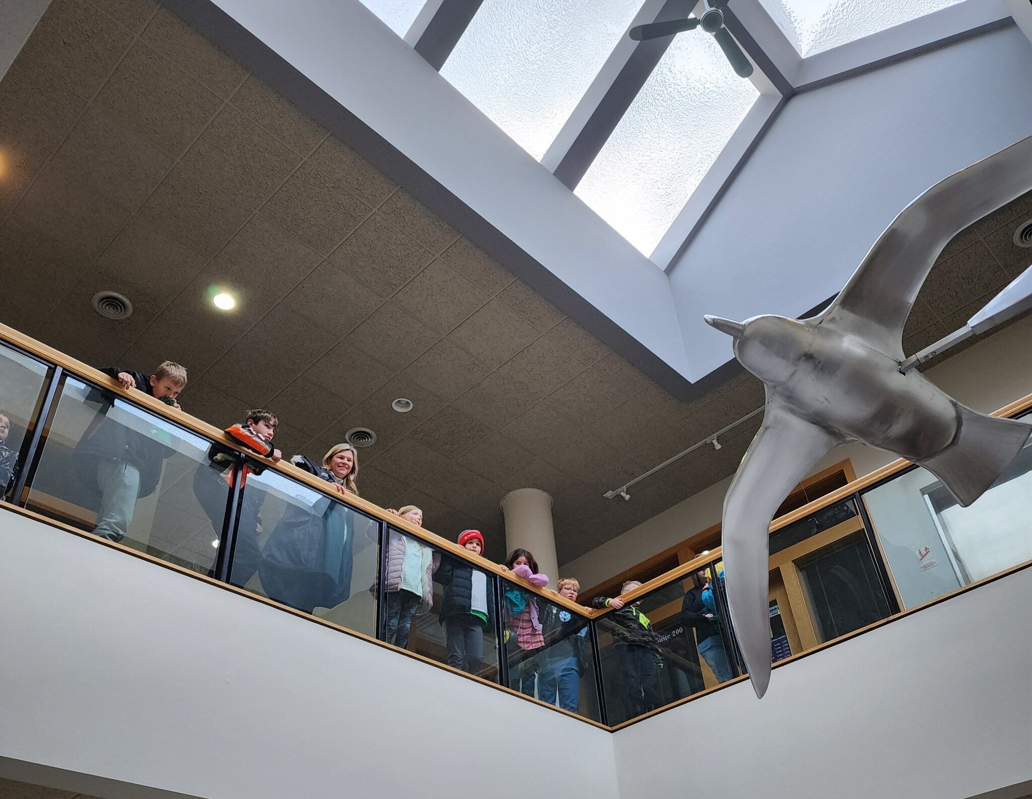 People look over a rail to view a large silver, metal bird sculpture inside a building under skylights.