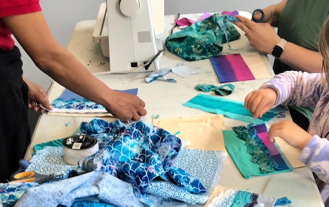 Three people work with fabric scraps on a white table with a sewing machine.