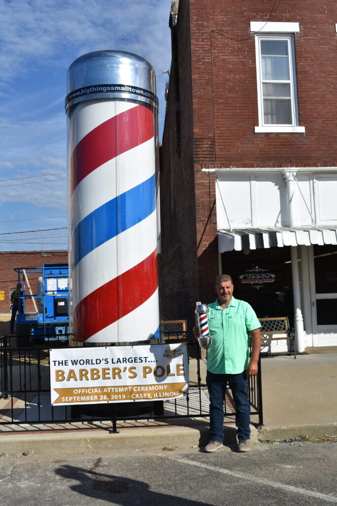 A man wearing a bright sea green shirt holds a barber's pole and stands next to a massive barber's pole.