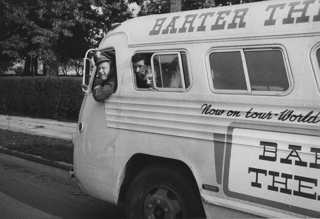 Three people sticking their heads out of the window of a bus, with signage reading, "Barter; now on tour"