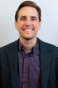 Headshot of a smiling person with light skin tone and short dark brown hair, wearing a blue blazer and checkered blue and red shirt.