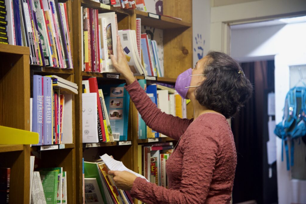 Person with light skin tone reaching up for a book on a bookshelf.
