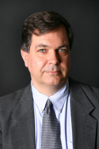 Headshot of male with light skin tone and brown hair wearing a grey suit and blue shirt