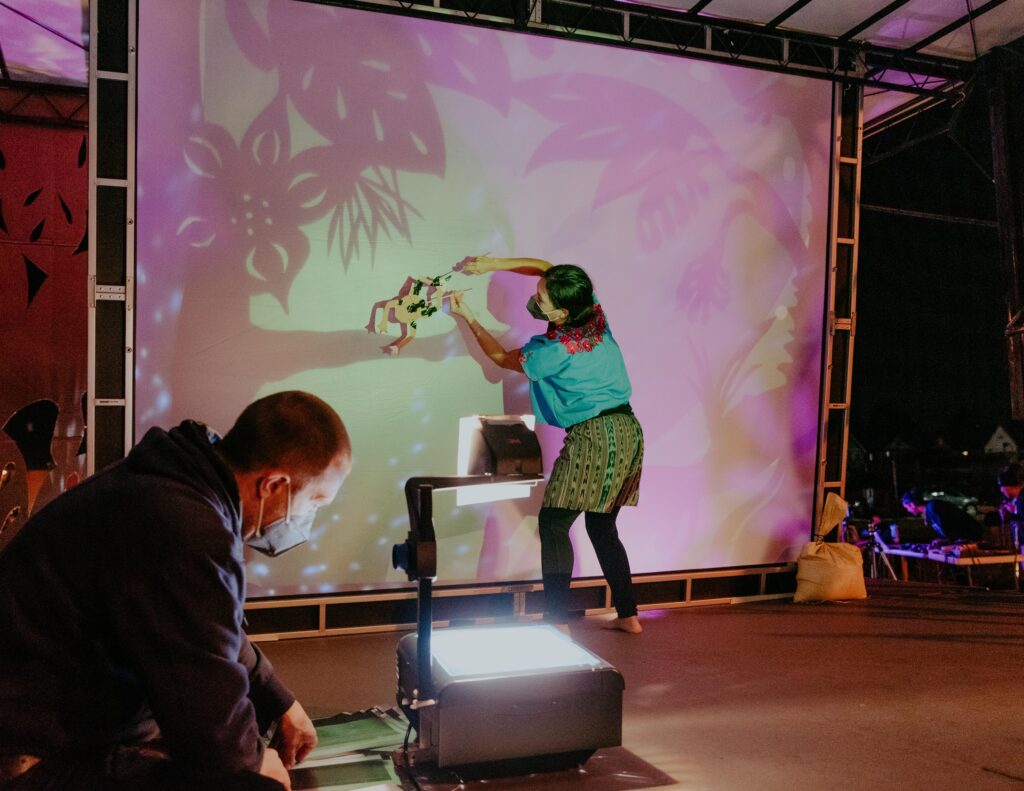 Star Girl Clan, a project led by artists Magdalena Kaluza and Rebekah Crisanta de Ybarra. A person holds a paper frog puppet up to a large screen, cast in purple and green with projections of branches and leaves.