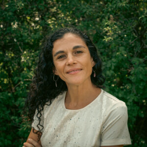 A woman with long, curly dark hair smiles softly at the camera. She is wearing a light-colored, short-sleeved shirt with subtle sequins and drop earrings. She stands in front of a lush green background of trees and leaves.