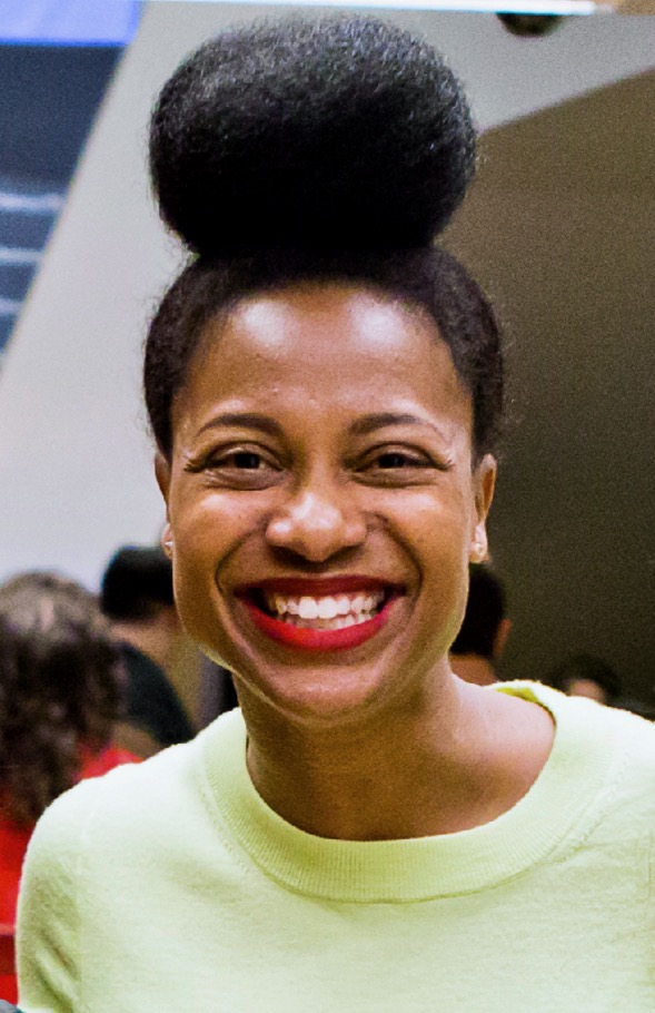 Headshot of a smiling person of medium dark skin tone, with black hair in a bun on top of their head, and wearing a bright yellow shirt.