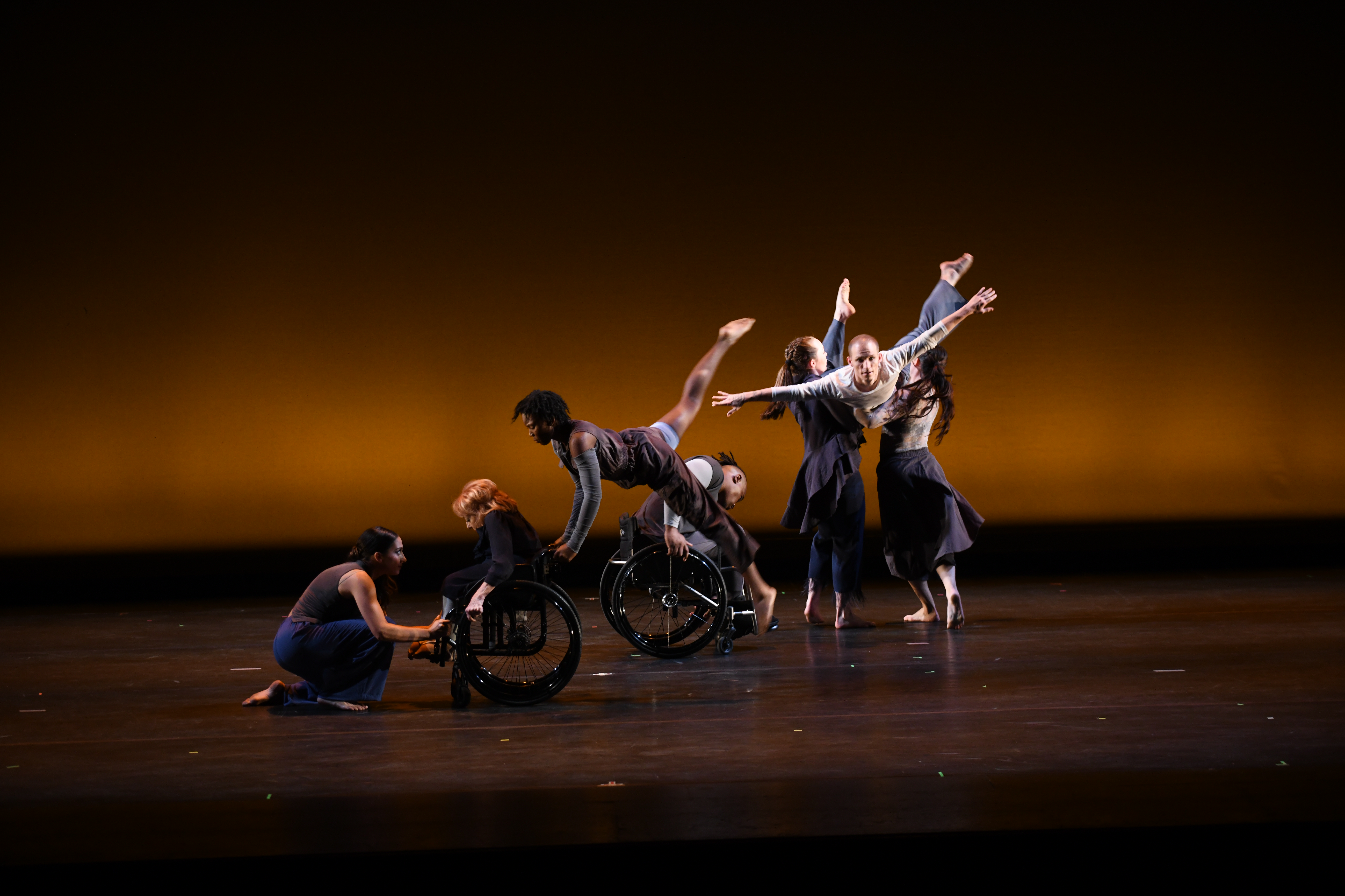 Dancers performing on stage with a moody lighting. Each dancer is in different pose with one lower to the ground and two in wheelchairs.