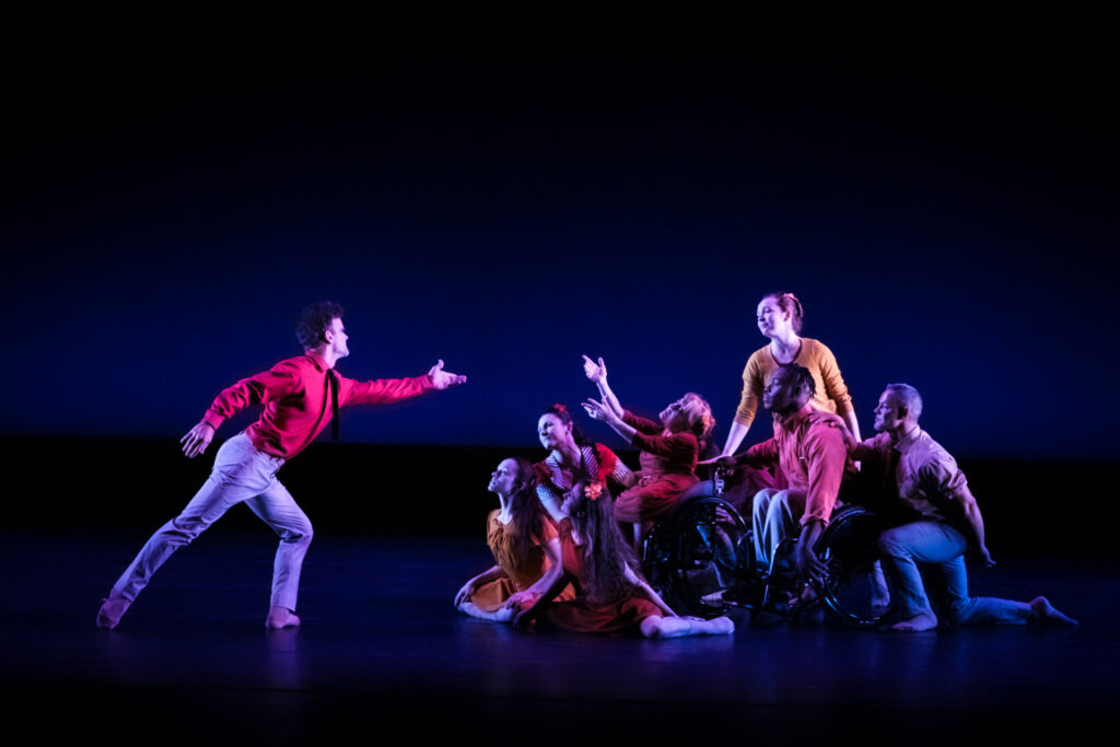 Dancers performing on stage with a moody lighting. Each dancer is in different pose with some sitting or lower to the ground, and two of them in wheelchairs.