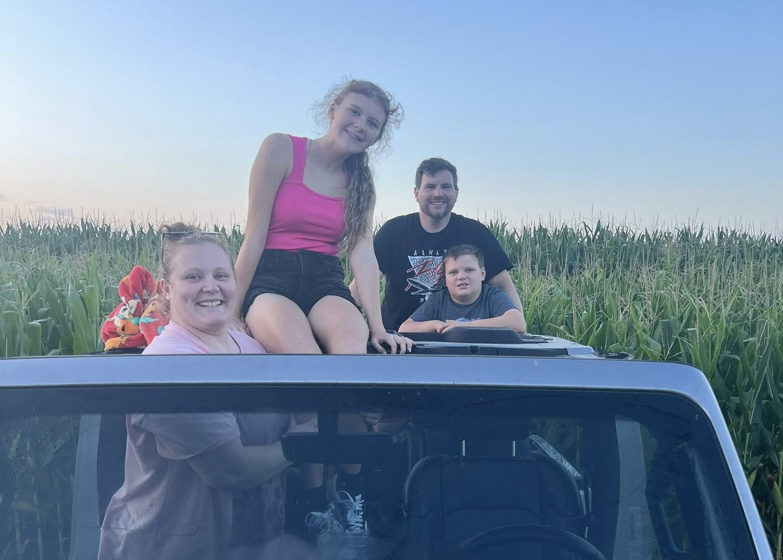 Four people siting on top of a car near a field and smiling.
