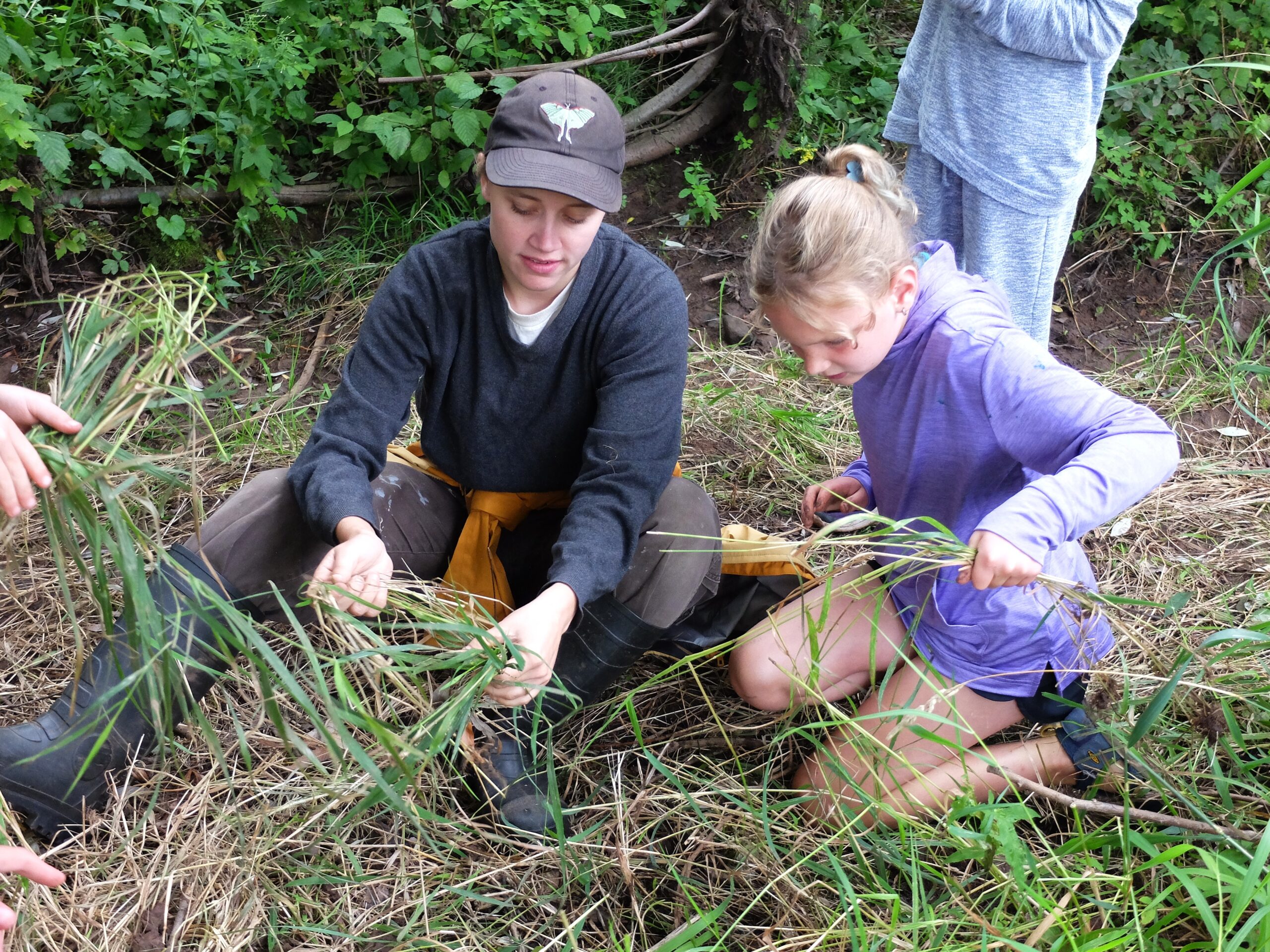 Two people with light skin tone sit in long grass and hold it.