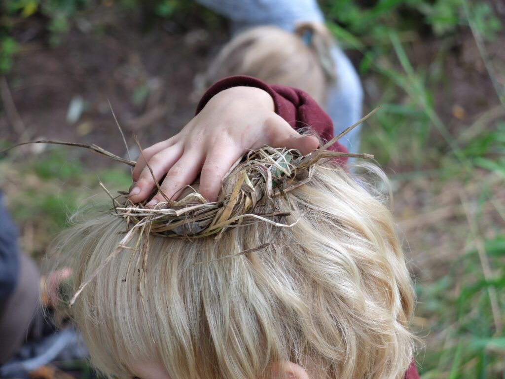 A child with blonde hair and light skin holds dried grass on their head.