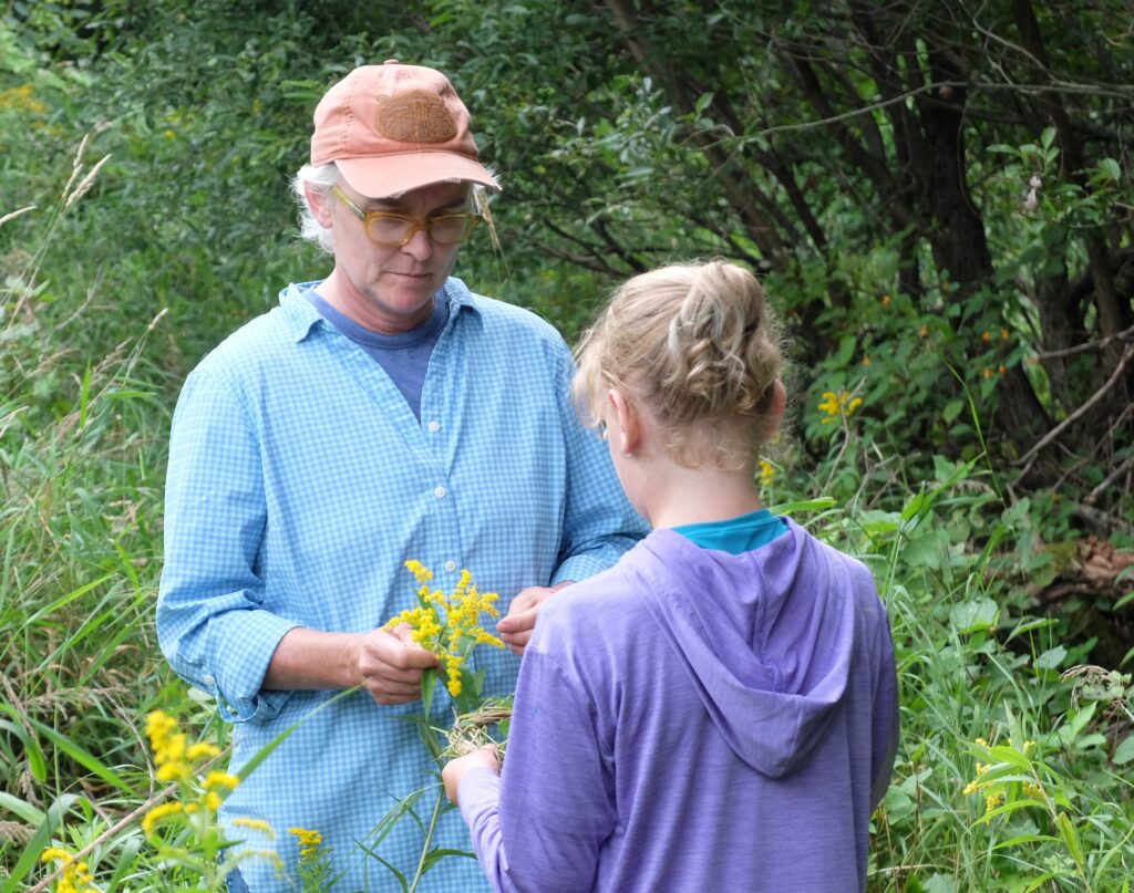 An adult with light skin holds yellow flowers in front of a child with blonde hair and light skin.