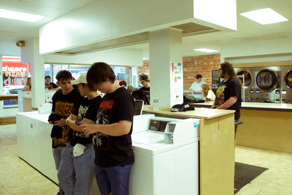 People standing around inside a laundromat. Most of them are wearing black tshirts and blue jeans.