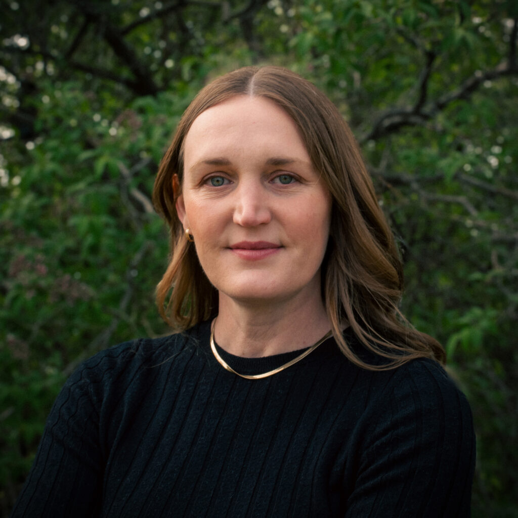 A woman with long, light brown hair gazes calmly at the camera. She is wearing a black, ribbed sweater and a gold necklace. The background is filled with dense, leafy green trees.