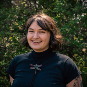 Headshot of a smiling person with light skin tone and dark brown hair, wearing a black shirt and dragonfly necklace.