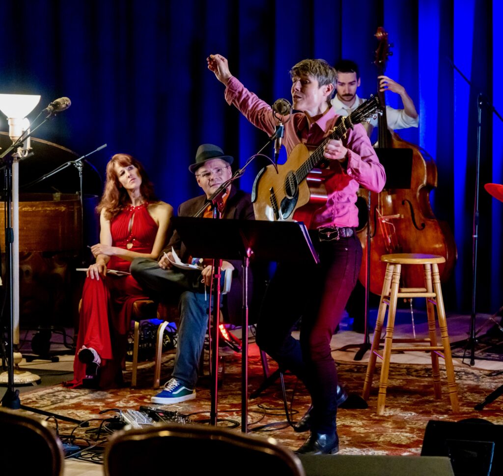 Ellis Delaney performs at the Red Jacket Jamboree in Calumet, MI. They are holding a guitar with one hand and another hand in the air, singing into a microphone. Behind them, two people sit and watch them, and another person plays standing bass.